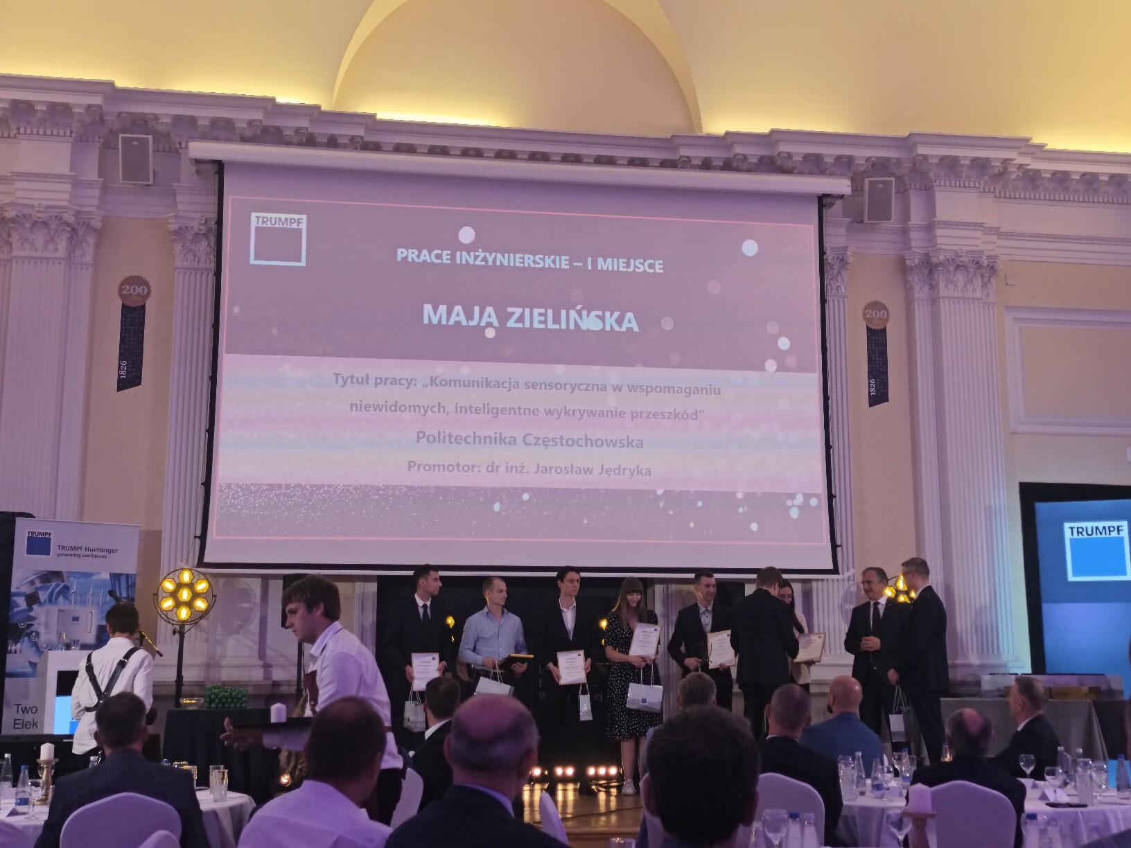 Zdjęcie z gali wręczenia nagród. Na scenie kilka osób odbiera dyplomy, a w tle wyświetlany jest slajd z napisem: ‘Prace inżynierskie – I miejsce, Maja Zielińska’ oraz tytułem pracy „Komunikacja sensoryczna w wspomaganiu niewidomych – inteligentne wykrywanie przeszkód”, Politechnika Częstochowska, promotor dr inż. Jarosław Jędryka. Na pierwszym planie widoczna jest publiczność siedząca przy stołach