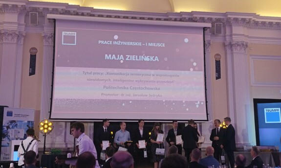 Zdjęcie z gali wręczenia nagród. Na scenie kilka osób odbiera dyplomy, a w tle wyświetlany jest slajd z napisem: ‘Prace inżynierskie – I miejsce, Maja Zielińska’ oraz tytułem pracy „Komunikacja sensoryczna w wspomaganiu niewidomych – inteligentne wykrywanie przeszkód”, Politechnika Częstochowska, promotor dr inż. Jarosław Jędryka. Na pierwszym planie widoczna jest publiczność siedząca przy stołach